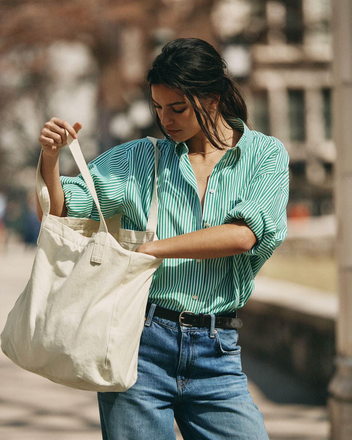 Women wearing green striped Gant shirt, blue jeans, black belt, carrying white tote bag in urban setting.